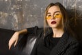 Young blonde woman in a black jacket and yellow glasses on a sofa against a gray wall Royalty Free Stock Photo