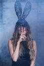 Young blonde woman in black rabbit or hare fancy mask and black dress. Finger on lips. Female on smoke and metal wall background Royalty Free Stock Photo