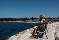 A young blonde woman in a blue dress sits on the bench near the sea and looks to the side. Royalty Free Stock Photo