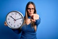 Young blonde woman with blue eyes holding big minute clock over isolated background pointing with finger to the camera and to you, Royalty Free Stock Photo