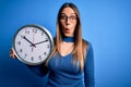 Young blonde woman with blue eyes holding big minute clock over isolated background scared in shock with a surprise face, afraid Royalty Free Stock Photo