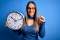 Young blonde woman with blue eyes holding big minute clock over isolated background surprised with an idea or question pointing Royalty Free Stock Photo