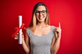 Young blonde woman with blue eyes wearing glasses holding graduated degree diploma surprised with an idea or question pointing Royalty Free Stock Photo