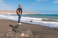 Young blonde woman with casual clothes and leather backpack standing on rock on windy beach