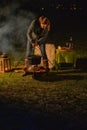 Young blonde woman is cooking soup in a black kettle outside on a winter evening. She is camping in the forest in a cozy