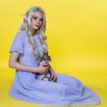 Young blonde woman cosplay elf sitting on yellow background, holding Sphinx kitten on her lap Royalty Free Stock Photo