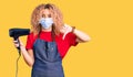 Young blonde woman with curly hair holding dryer blow wearing safety mask for coranvirus with angry face, negative sign showing