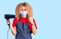 Young blonde woman with curly hair holding dryer blow wearing safety mask for coranvirus annoyed and frustrated shouting with