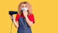 Young blonde woman with curly hair holding dryer blow wearing safety mask for coranvirus covering mouth with hand, shocked and