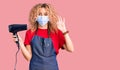 Young blonde woman with curly hair holding dryer blow wearing safety mask for coranvirus doing ok sign with fingers, smiling