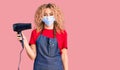 Young blonde woman with curly hair holding dryer blow wearing safety mask for coranvirus looking positive and happy standing and