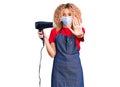 Young blonde woman with curly hair holding dryer blow wearing safety mask for coranvirus with open hand doing stop sign with