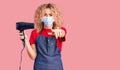 Young blonde woman with curly hair holding dryer blow wearing safety mask for coranvirus pointing with finger to the camera and to