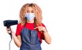 Young blonde woman with curly hair holding dryer blow wearing safety mask for coranvirus pointing finger to one self smiling happy