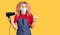 Young blonde woman with curly hair holding dryer blow wearing safety mask for coranvirus pointing thumb up to the side smiling