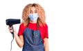 Young blonde woman with curly hair holding dryer blow wearing safety mask for coranvirus scared and amazed with open mouth for