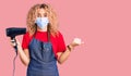Young blonde woman with curly hair holding dryer blow wearing safety mask for coranvirus screaming proud, celebrating victory and