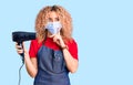 Young blonde woman with curly hair holding dryer blow wearing safety mask for coranvirus serious face thinking about question with