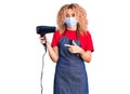 Young blonde woman with curly hair holding dryer blow wearing safety mask for coranvirus smiling happy pointing with hand and