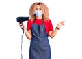 Young blonde woman with curly hair holding dryer blow wearing safety mask for coranvirus smiling happy pointing with hand and