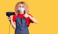 Young blonde woman with curly hair holding dryer blow wearing safety mask for coranvirus smiling happy and positive, thumb up