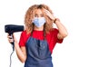 Young blonde woman with curly hair holding dryer blow wearing safety mask for coranvirus stressed and frustrated with hand on