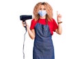 Young blonde woman with curly hair holding dryer blow wearing safety mask for coranvirus surprised with an idea or question