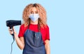Young blonde woman with curly hair holding dryer blow wearing safety mask for coranvirus thinking attitude and sober expression