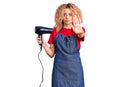 Young blonde woman with curly hair wearing hairdresser apron and holding dryer blow with open hand doing stop sign with serious