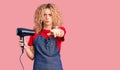 Young blonde woman with curly hair wearing hairdresser apron and holding dryer blow pointing with finger to the camera and to you,