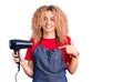 Young blonde woman with curly hair wearing hairdresser apron and holding dryer blow pointing finger to one self smiling happy and