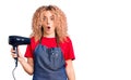 Young blonde woman with curly hair wearing hairdresser apron and holding dryer blow scared and amazed with open mouth for