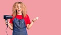 Young blonde woman with curly hair wearing hairdresser apron and holding dryer blow screaming proud, celebrating victory and Royalty Free Stock Photo