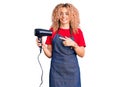 Young blonde woman with curly hair wearing hairdresser apron and holding dryer blow smiling happy pointing with hand and finger