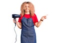 Young blonde woman with curly hair wearing hairdresser apron and holding dryer blow smiling happy pointing with hand and finger to