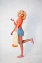 a young blonde woman with a curly hairstyle in an orange blouse and denim shorts holds a mesh bag with oranges