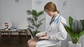 Young blonde woman doctor using laptop sitting on chair at clinic waiting room Royalty Free Stock Photo
