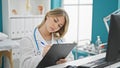 Young blonde woman doctor writing on clipboard thinking at clinic Royalty Free Stock Photo