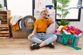 Young blonde woman doing laundry sitting by washing machine surprised with hand on head for mistake, remember error