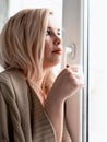 Young blonde woman is dreaming about something while sitting on the window-sill. she holding a tea cup and drinking Royalty Free Stock Photo