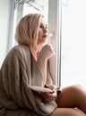 Young blonde woman is dreaming about something while sitting on the window-sill. she holding a tea cup and drinking Royalty Free Stock Photo