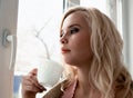 Young blonde woman is dreaming about something while sitting on the window-sill. she holding a tea cup and drinking Royalty Free Stock Photo