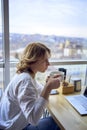 young blonde woman drinking coffee and working on a laptop in a cafe with panoramic windows and a view of the city from above Royalty Free Stock Photo