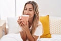 Young blonde woman drinking cup of coffee sitting on bed at bedroom Royalty Free Stock Photo