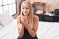 Young blonde woman drinking cup of coffee sitting on bed at bedroom Royalty Free Stock Photo