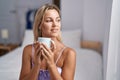 Young blonde woman drinking cup of coffee sitting on bed at bedroom Royalty Free Stock Photo