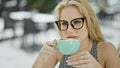 Young blonde woman drinking cup of coffee sitting on table thinking at coffee shop terrace Royalty Free Stock Photo