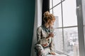 Young blonde woman drinking tea, coffee and looking through big window, happy, good morning at home. Wearing silk Royalty Free Stock Photo