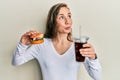 Young blonde woman eating a tasty classic burger with fries and soda smiling looking to the side and staring away thinking Royalty Free Stock Photo