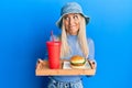Young blonde woman eating a tasty classic burger and soda smiling looking to the side and staring away thinking Royalty Free Stock Photo